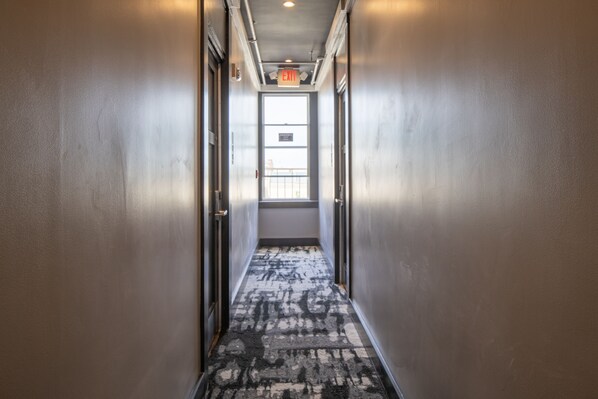A narrow hallway with a patterned carpet, metal walls, and an exit sign. - Hotel 32One (San Francisco)
