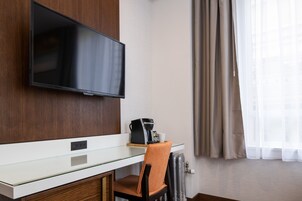 A hotel room with a flat-screen TV mounted on the wall, a coffee machine on a glass table, a wooden chair, and a window with curtains.