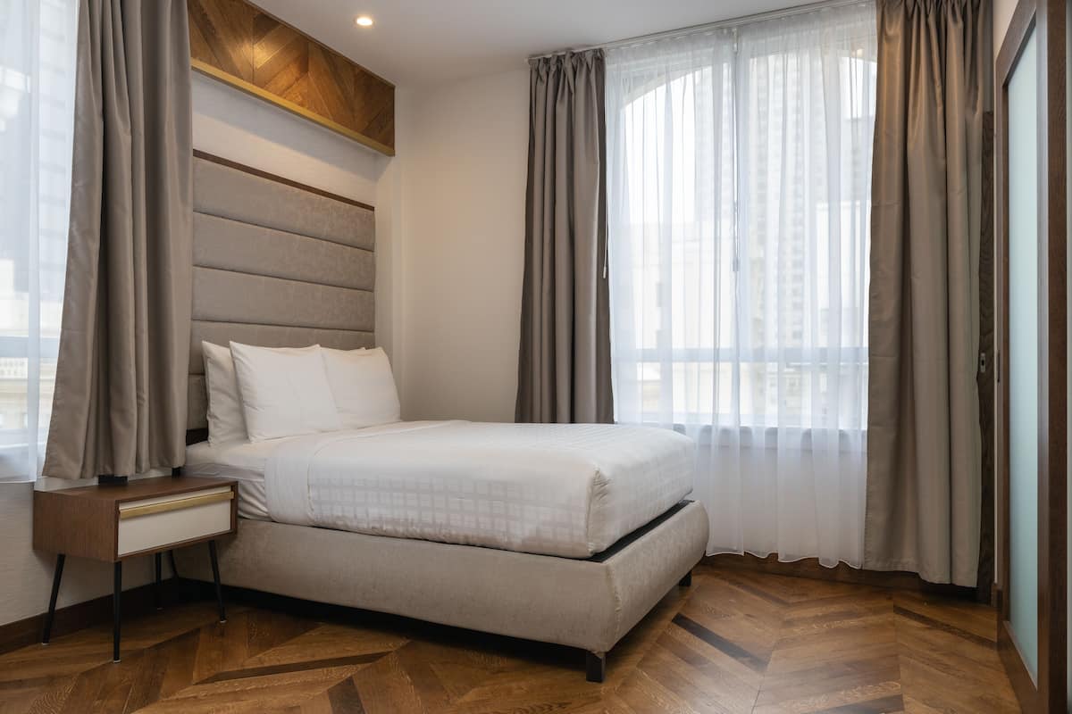 Habitación de hotel moderna con una cama grande, una mesita de noche y vistas de la ciudad a través de cortinas translúcidas.