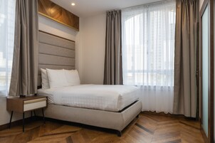 A modern hotel room with a large bed, a nightstand, and a view of the city through sheer curtains.