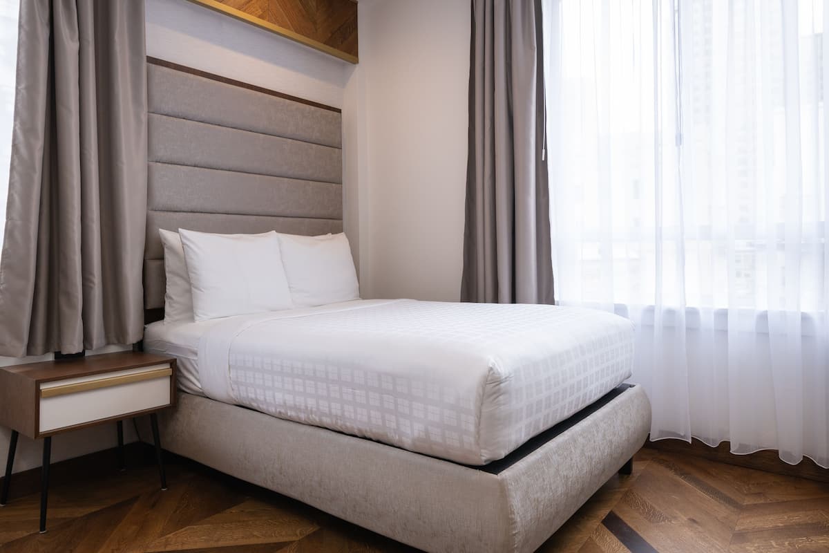 Una cama bien tendida con cabecera, una mesita de noche con cajón y una ventana con cortinas traslúcidas.