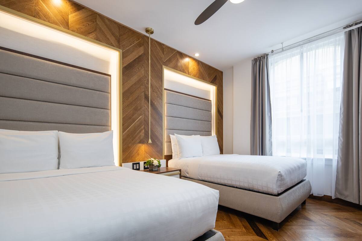 Habitación de hotel con dos camas, una pared decorativa de madera, un ventilador de techo y un ventanal con cortinas.