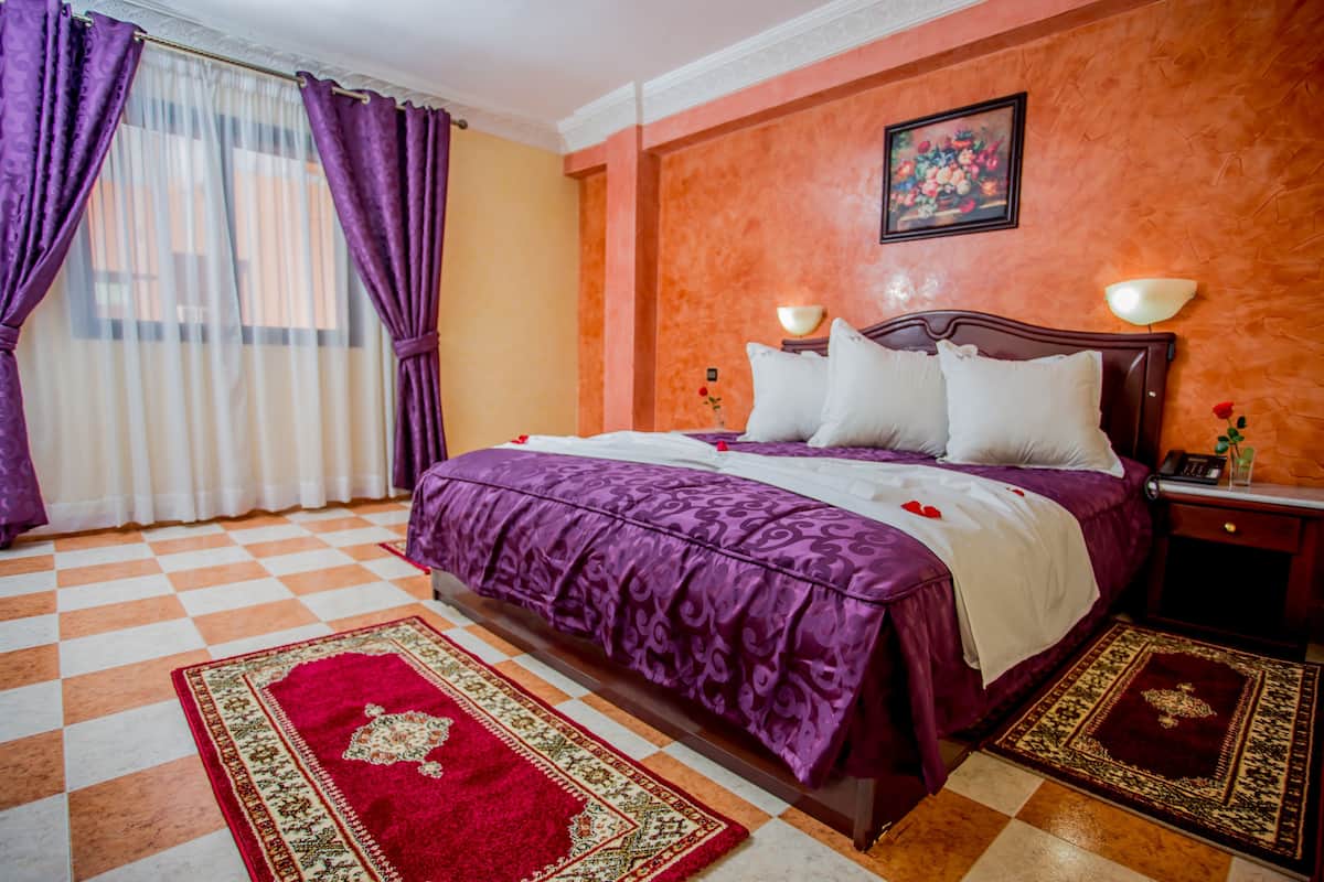 Habitación de hotel con piso a cuadros, una cama grande con cubrecama morado, almohadas blancas y un tapete rojo y dorado.
