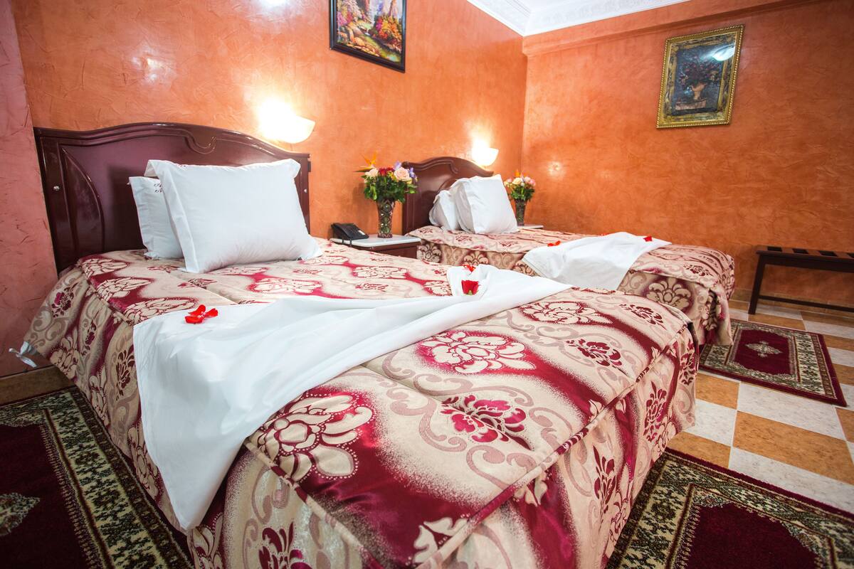 Habitación de hotel con dos camas, cada una con cubrecama estampado con motivos florales, cabecero de madera y mesita de noche con un jarrón de flores.