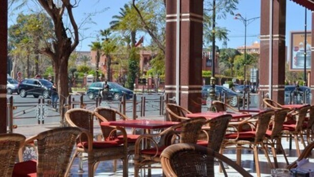 An outdoor cafe with wicker chairs and red tables, set against a backdrop of palm trees and a clear sky.