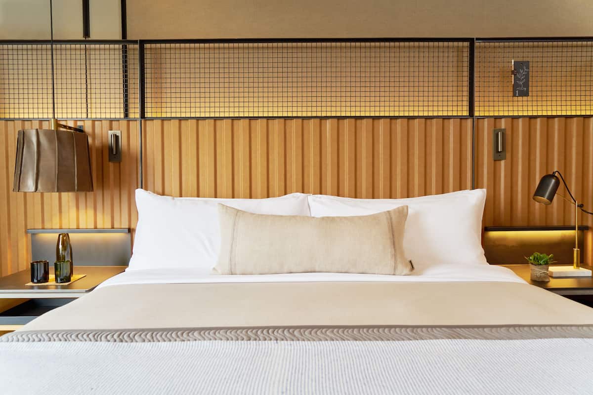 A neatly made bed with white linens and a beige pillow, a bedside lamp, and a small plant on a bedside table.