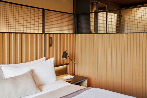 Habitación de hotel con cama, mesita de noche, lámpara y vistas a través de un panel de madera.