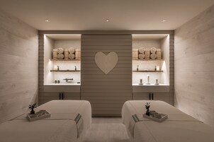 Egy modern hotelszoba, visszafogott színvilággal, két ággyal, központi, szív alakú díszítéssel, valamint beépített polcokkal, melyeken dekoratív tárgyak találhatók.
