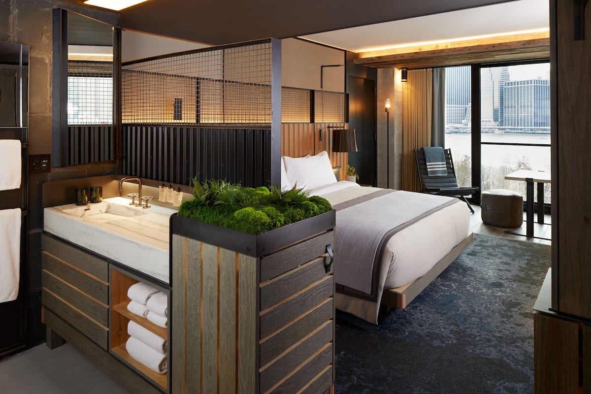 A modern hotel room with a large bed, a built-in bathtub with a green moss feature, a separate seating area with a table and chair, and a city view through large windows.