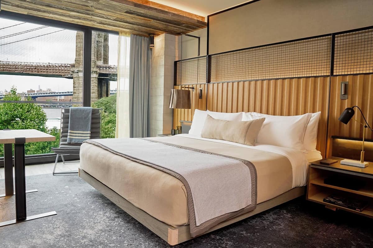A modern hotel room with a large bed, a small table, a chair, and a view of a bridge and greenery.