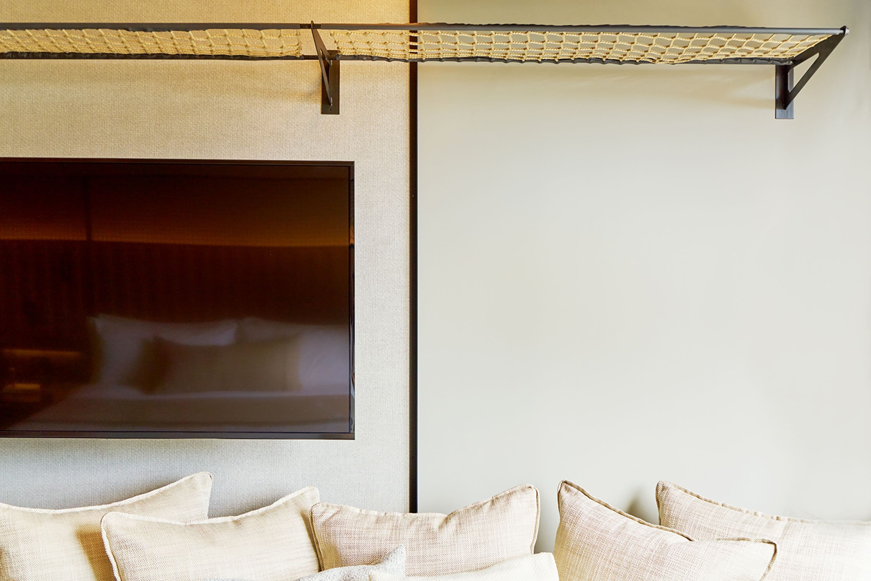 A modern living room with a large flat-screen TV mounted on the wall, a sofa with multiple cushions, and a ceiling-mounted netting structure.