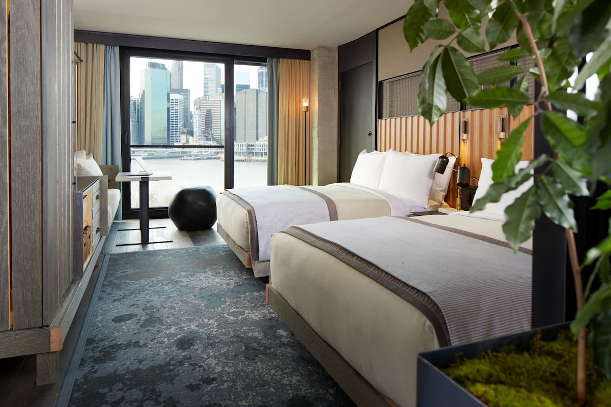 A hotel room with two beds, a view of the city, and a small table with a chair.