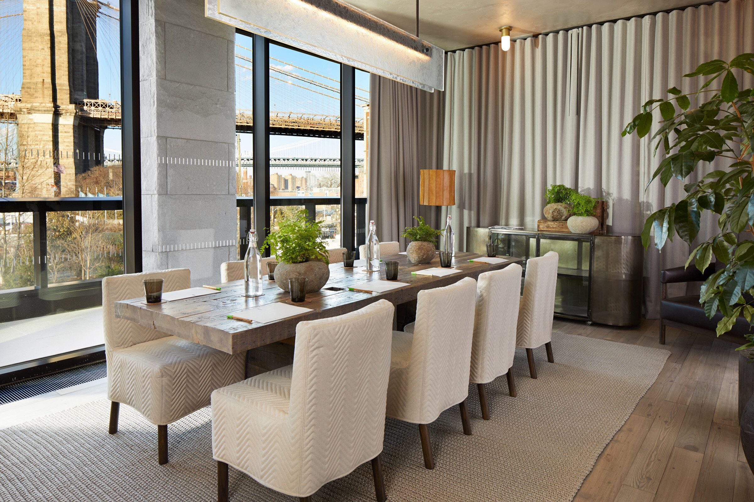 A modern dining area with a large table, white upholstered chairs, and a view of a bridge.