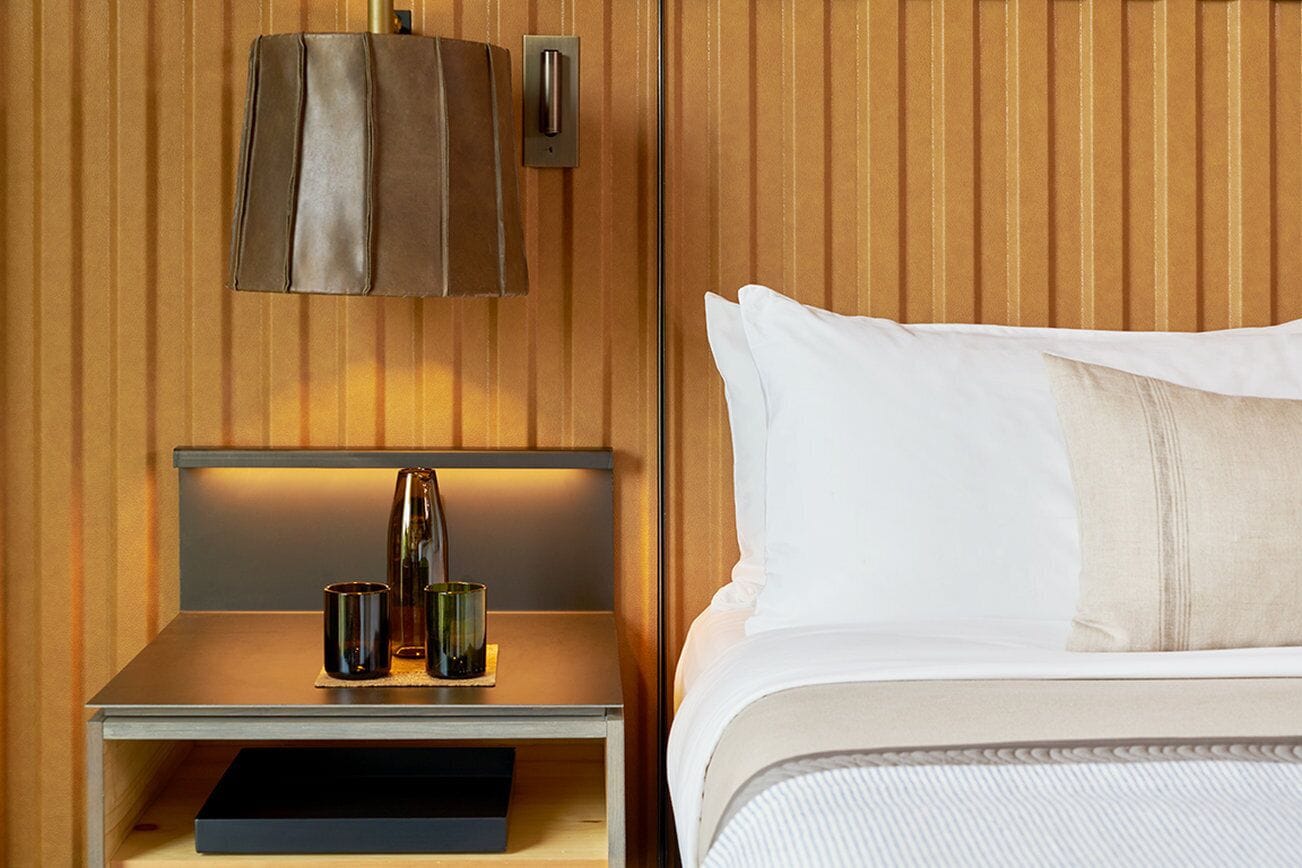 A neatly made bed with white linens and a single pillow, a bedside table with a lamp and two glasses, and a wall-mounted light fixture.