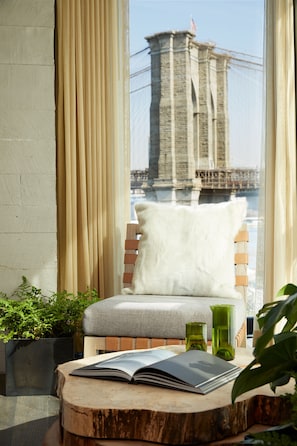 A room with a view of the Brooklyn Bridge, a wooden chair with a white cushion, a book on a wooden table, and green glass vases.