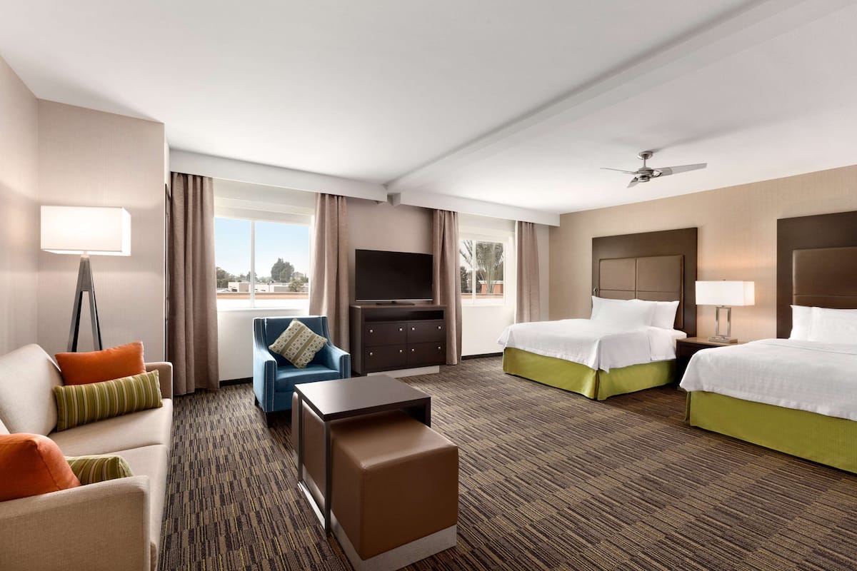 A hotel room with a sofa, a chair, a desk, a TV, a bed, and a ceiling fan.