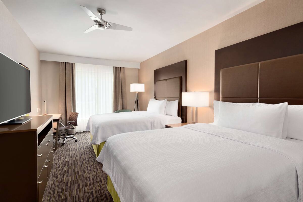 A hotel room with two beds, a desk, a chair, a television, and a ceiling fan.