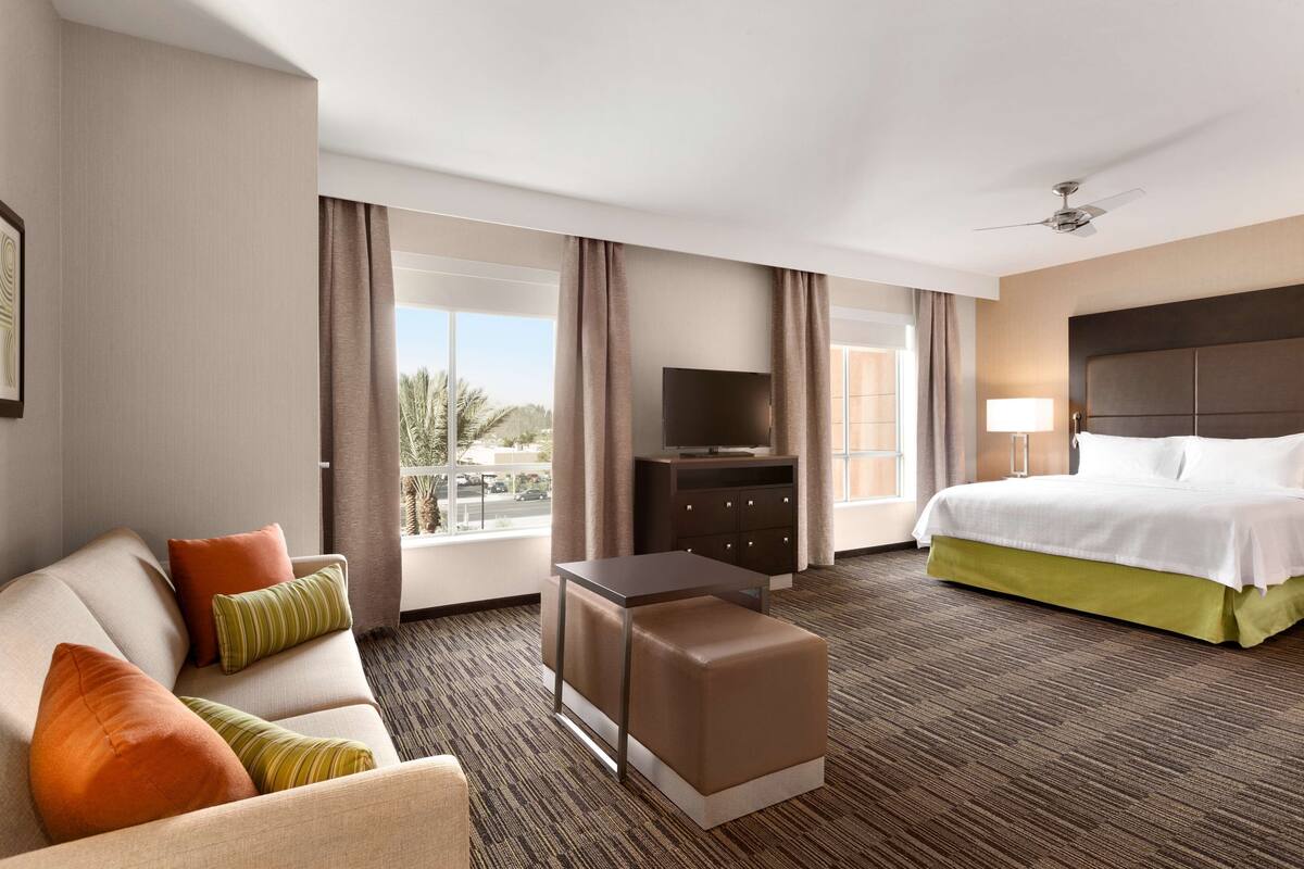 A hotel room with a sofa, ottoman, TV, bed, and a view of palm trees.