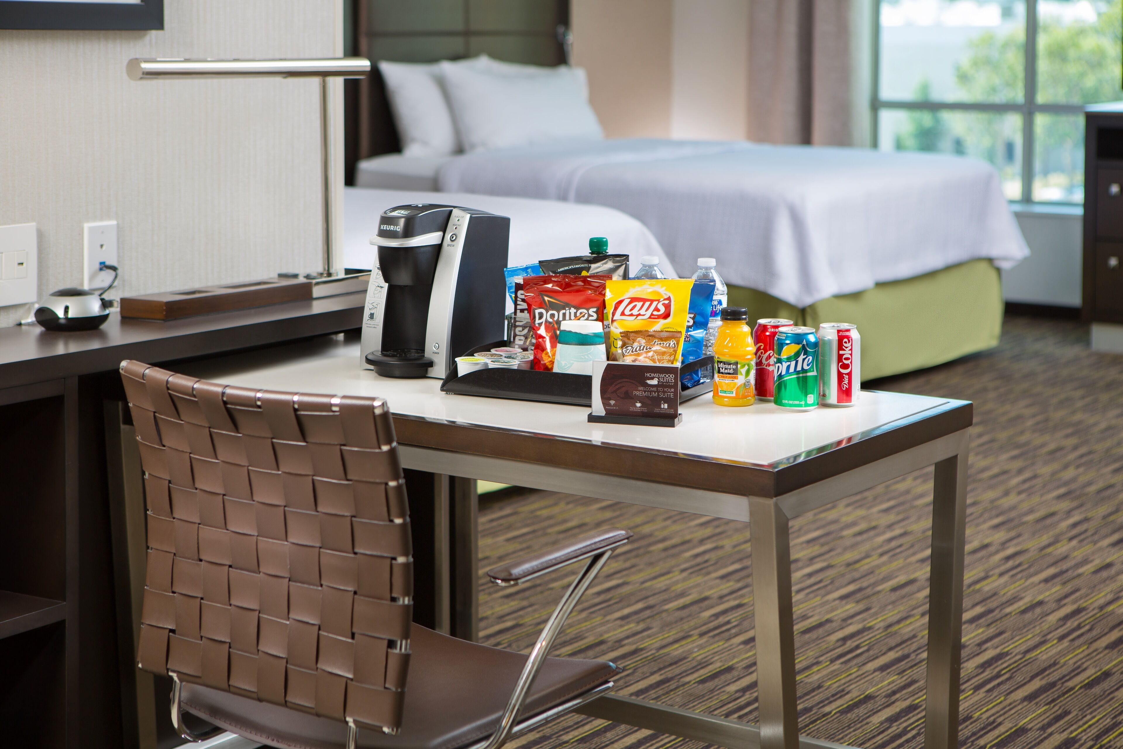 A hotel room with a bed, a desk with a coffee maker, snacks, and a chair.
