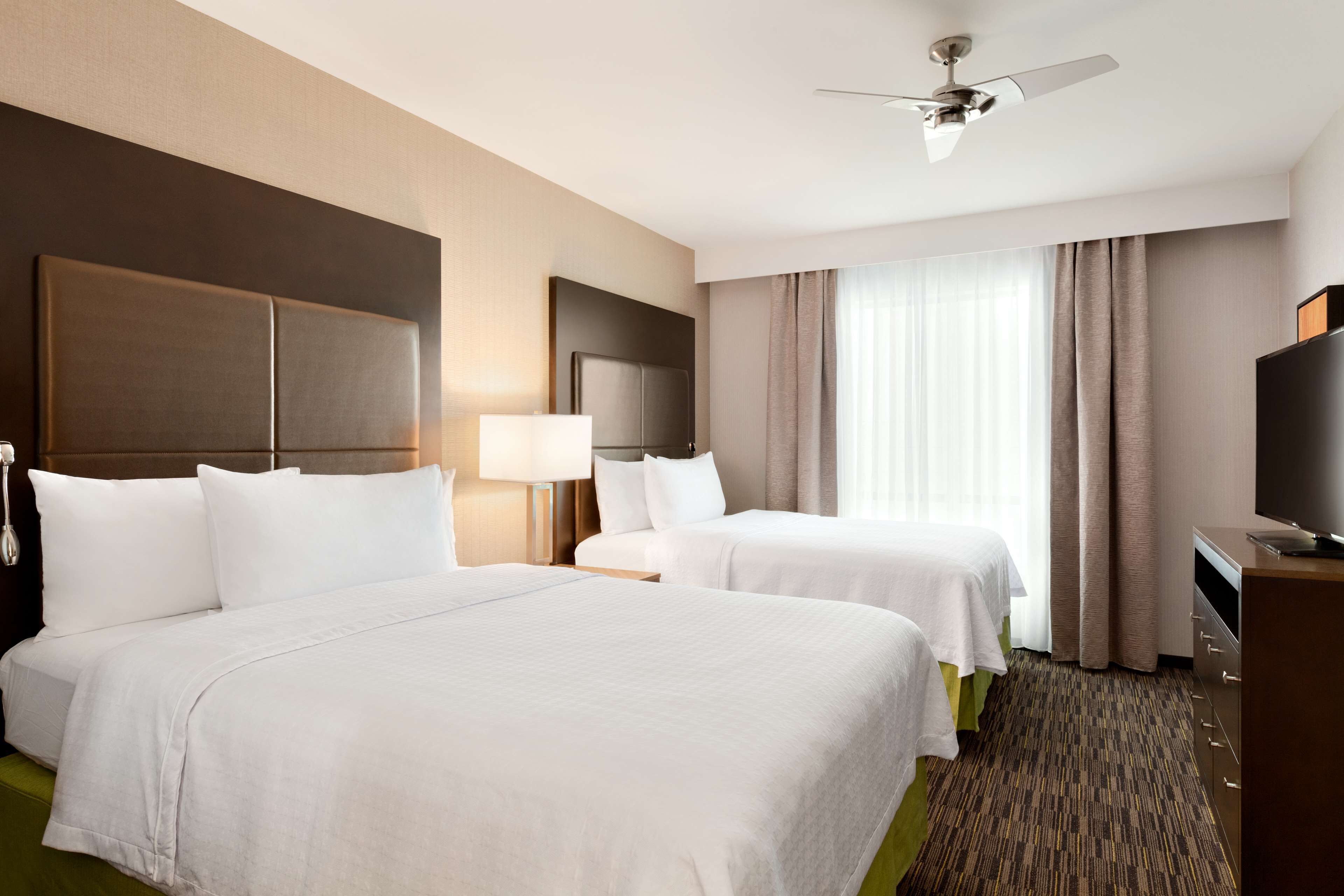 A hotel room with two beds, a desk, a television, and a ceiling fan.