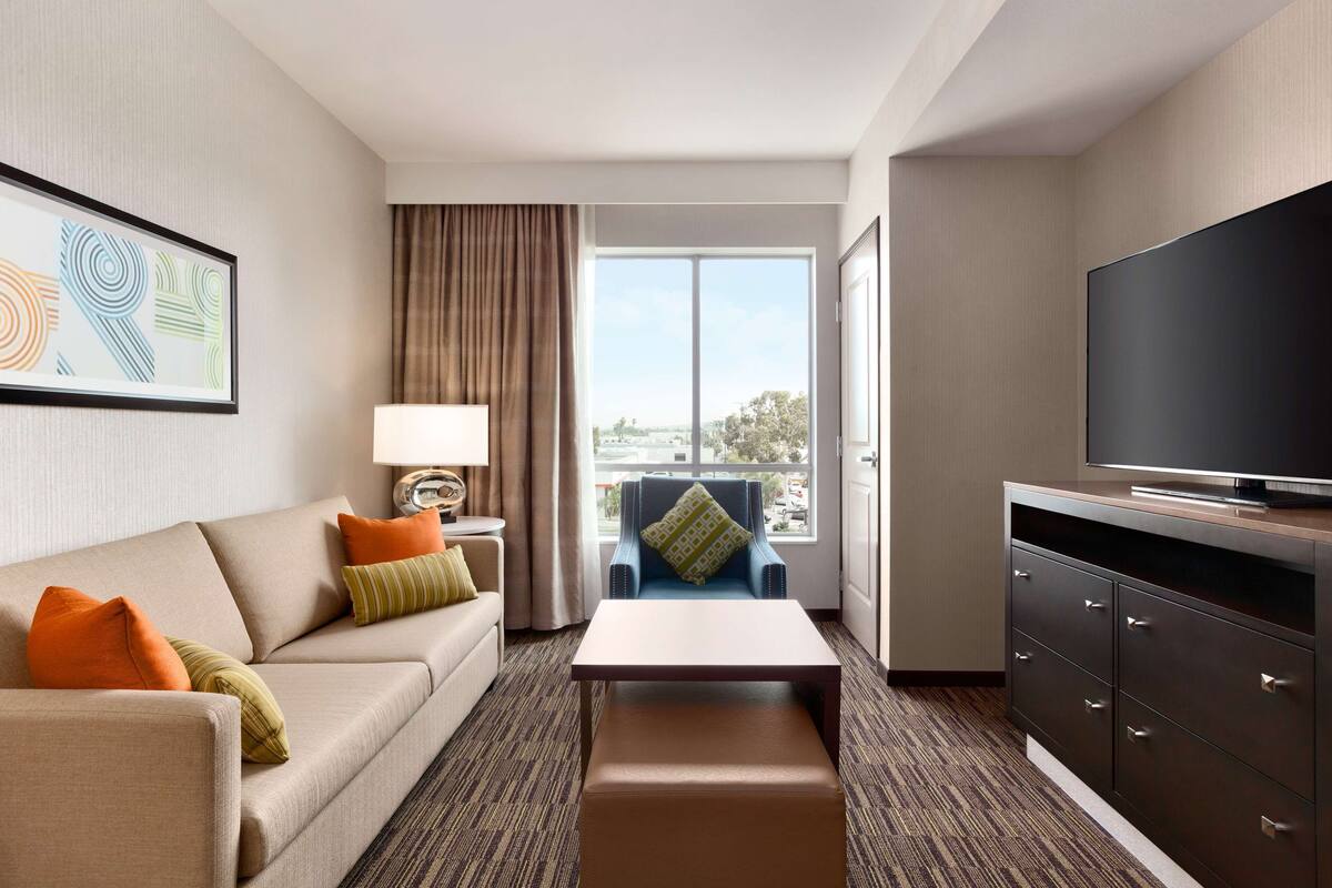 A hotel room with a sofa, a coffee table, a TV, a chair, and a window with a view.