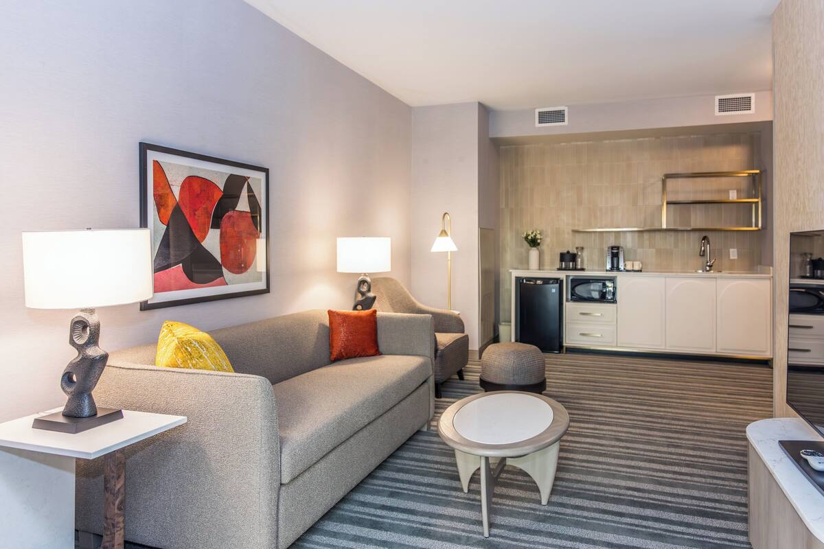 Une chambre d’hôtel moderne avec un canapé gris, une table basse ronde, une petite table blanche, une lampe et un coin cuisine équipé d’un four à micro-ondes et d’une machine à café.