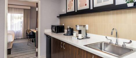 A modern kitchen with a stainless steel sink, a microwave, and a coffee maker. There is a dining area with a chair and a table.