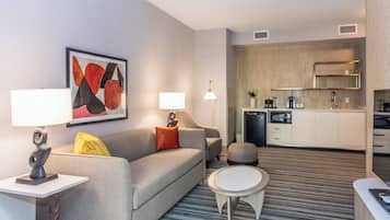 A modern hotel room with a grey sofa, a round coffee table, a small white table, a lamp, and a kitchen area with a microwave and a coffee maker.