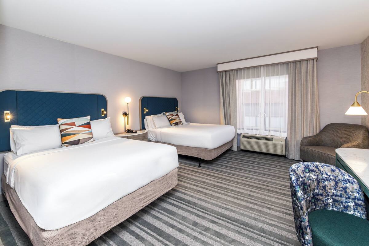 Une chambre d’hôtel avec deux lits, un bureau, une chaise, une grande fenêtre avec des rideaux et un radiateur fixé au mur.