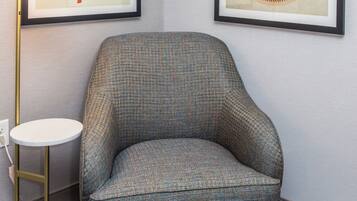 A corner room with a patterned armchair, a round side table, a floor lamp, and two framed artworks on the wall.