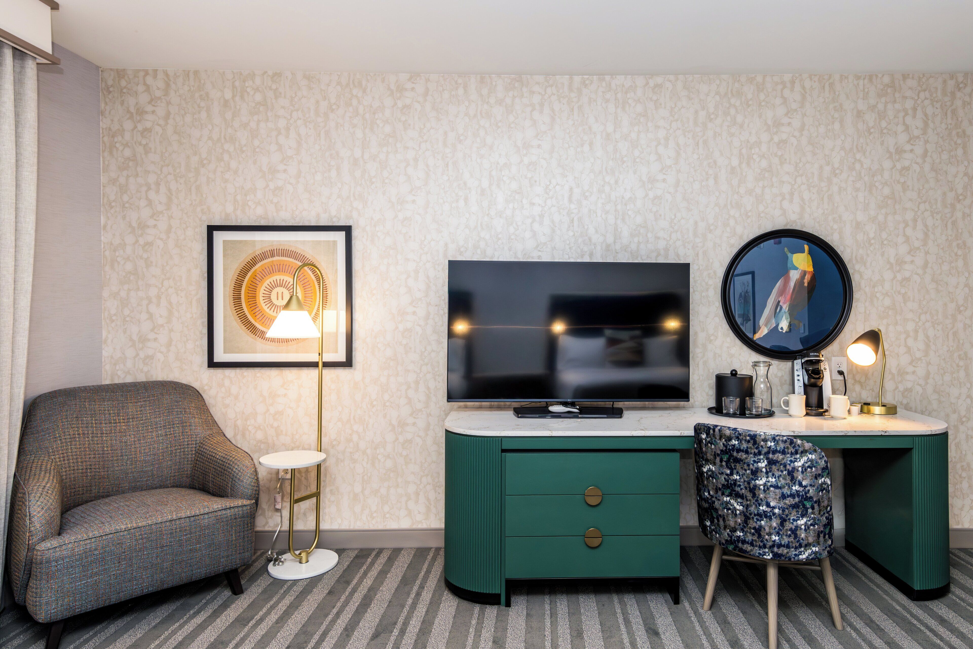Habitación de hotel con un escritorio verde, televisor, una obra de arte enmarcada, una silla y una lámpara de pie.
