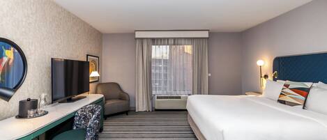A hotel room with a bed, a desk with a chair, a TV, and a window with curtains.