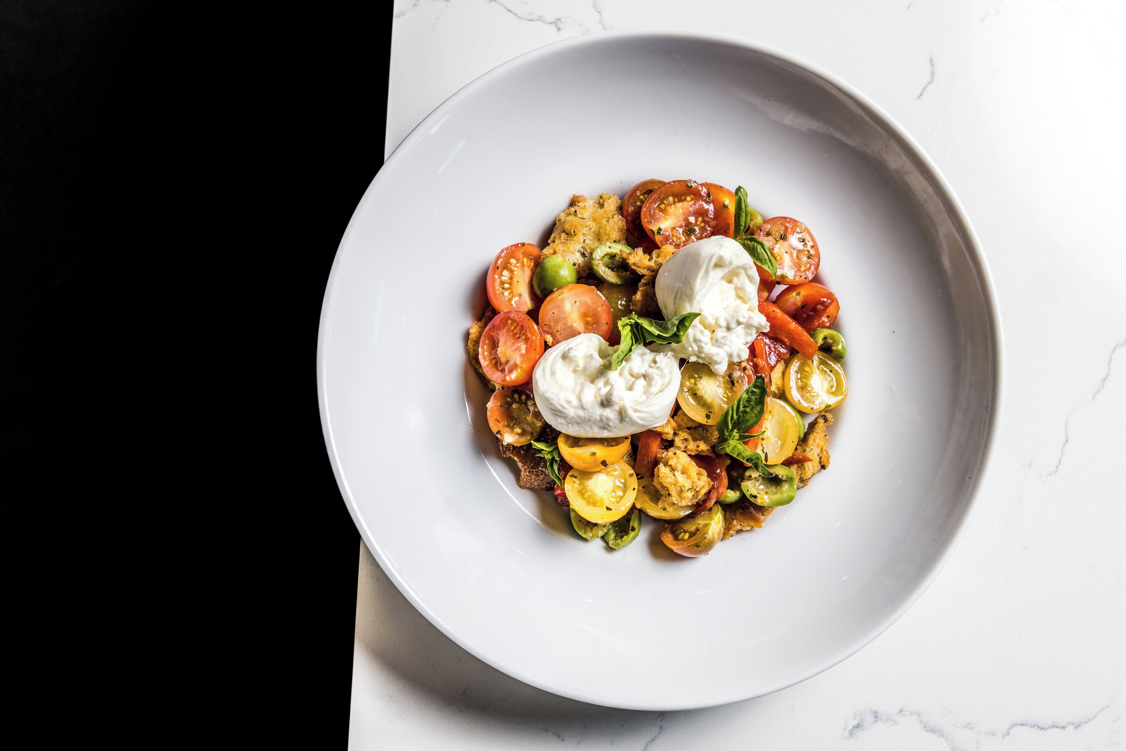 Un plato blanco con una ensalada de tomates, verduras y mozzarella sobre una superficie de mármol.