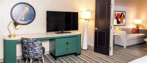 A hotel room with a TV, a desk with a chair, a lamp, a door, and a couch.
