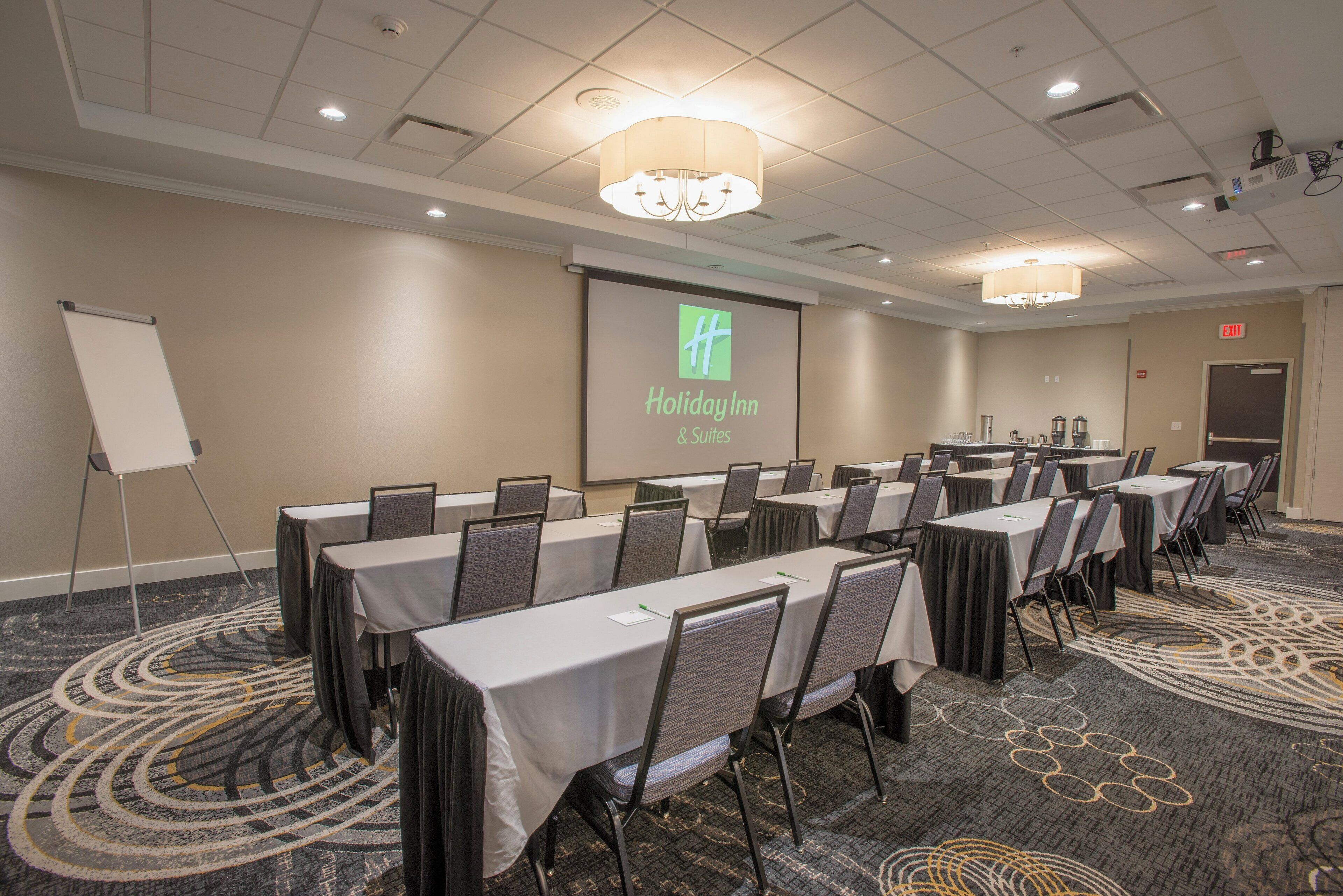 A conference room with tables and chairs, a projector screen displaying a logo, and an exit sign.