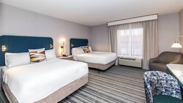 Ein Hotelzimmer mit zwei Betten, einem Schreibtisch, einem Sessel, einem großen Fenster mit Vorhängen und einem Wandheizkörper.