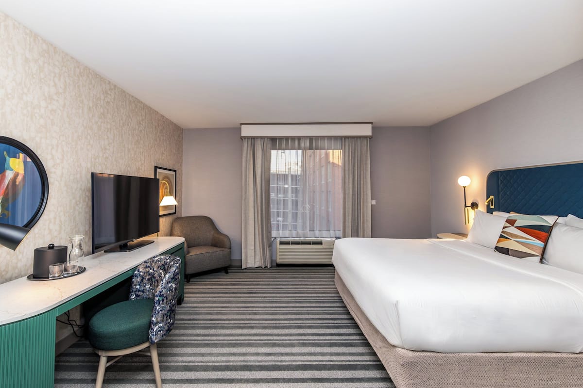 Une chambre d’hôtel comprenant un lit, un bureau avec une chaise, une télévision et une fenêtre avec des rideaux.