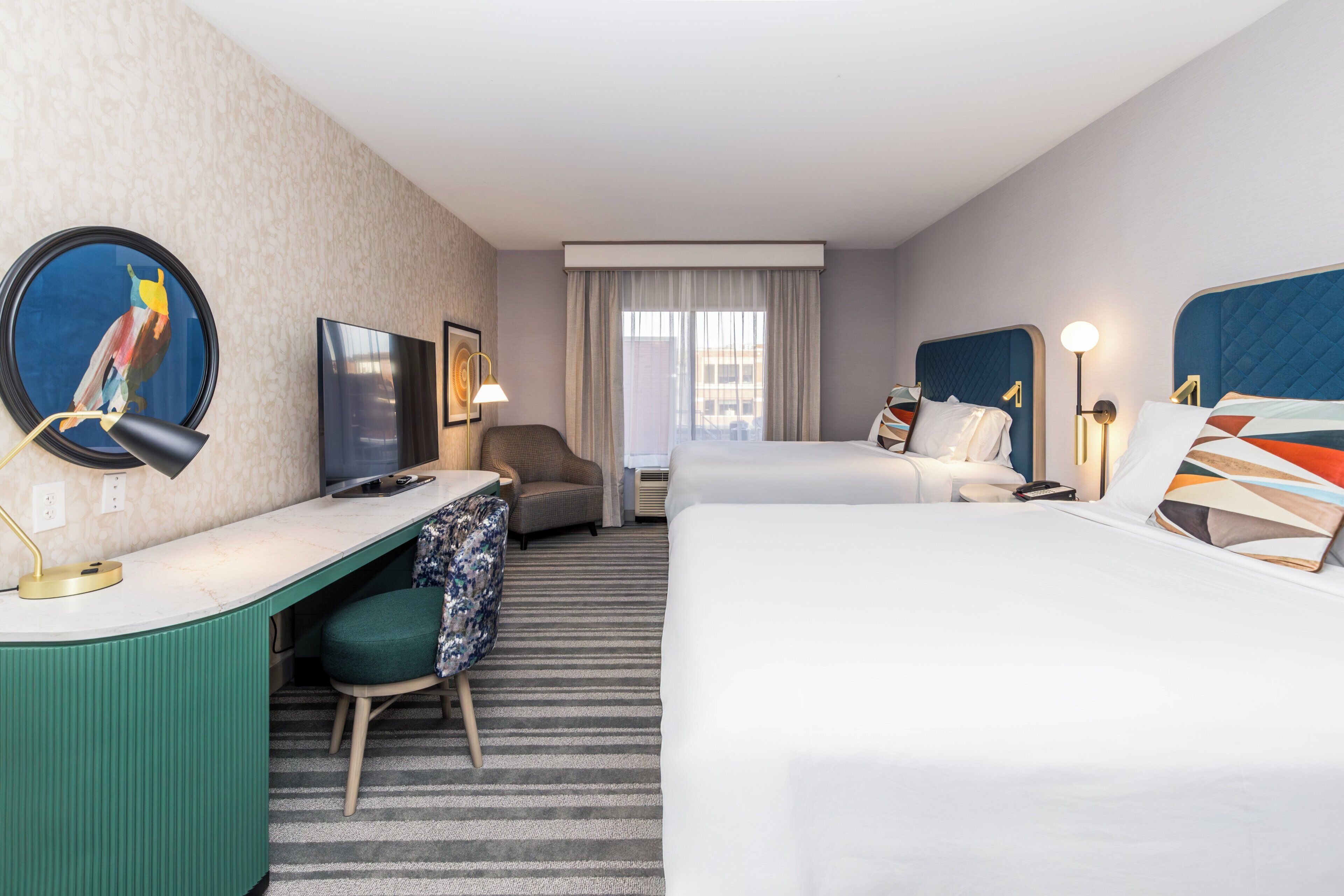 A hotel room with a bed, a desk with a computer, a chair, a TV, and a window with curtains.