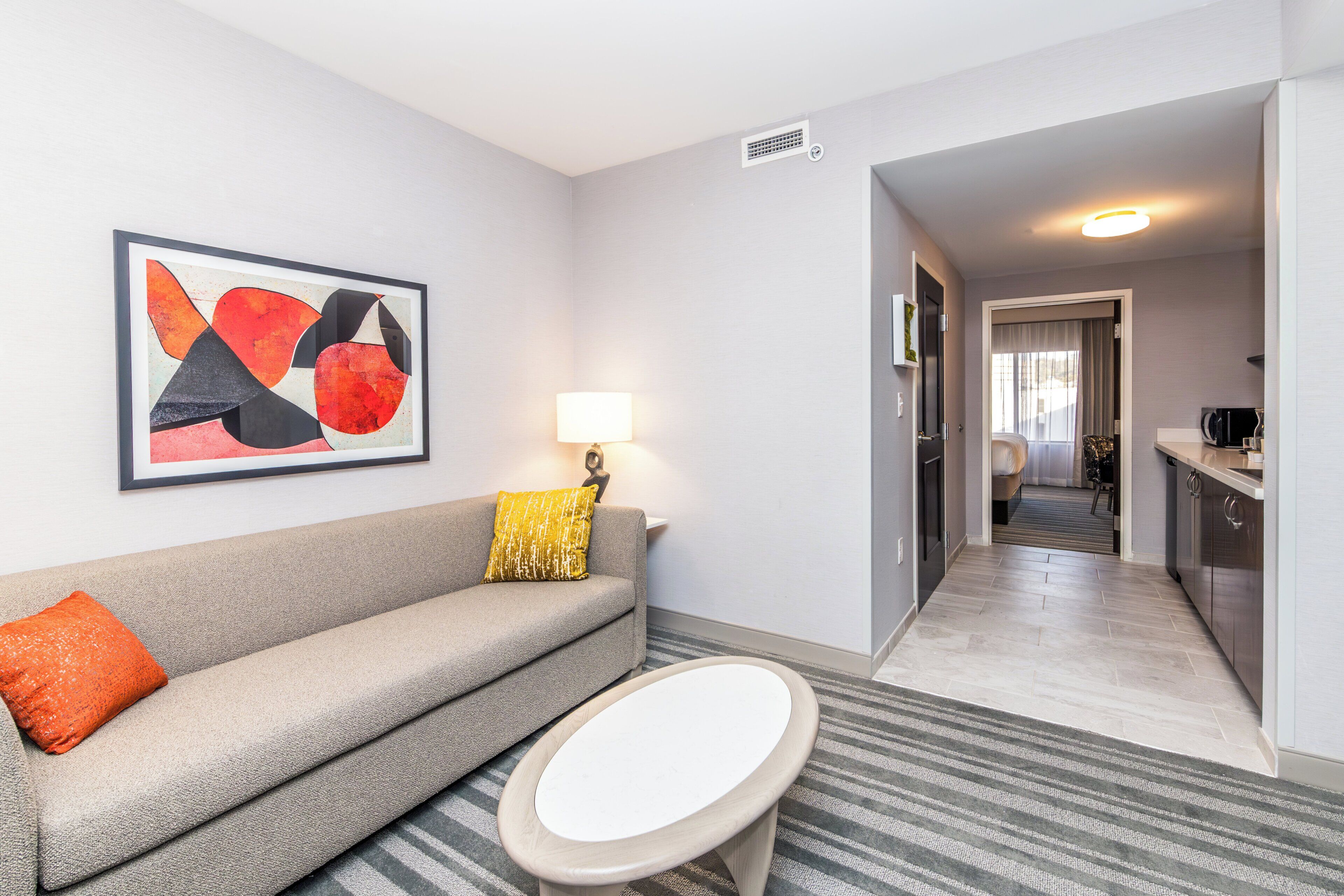 A modern hotel room with a grey sofa, a small round table, a framed abstract painting, a lamp, and a view into the kitchen and dining area.