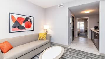 A modern hotel room with a grey sofa, a small round table, a framed abstract painting, a lamp, and a view into the kitchen and dining area.