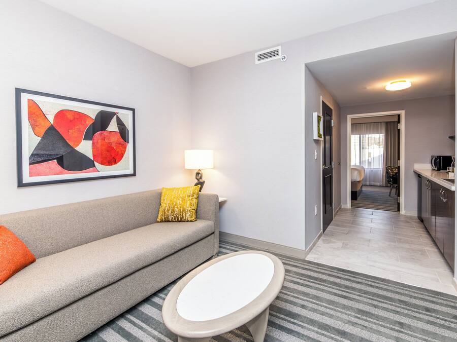 A modern hotel room with a grey sofa, a small round table, a framed abstract painting, a lamp, and a view into the kitchen and dining area.