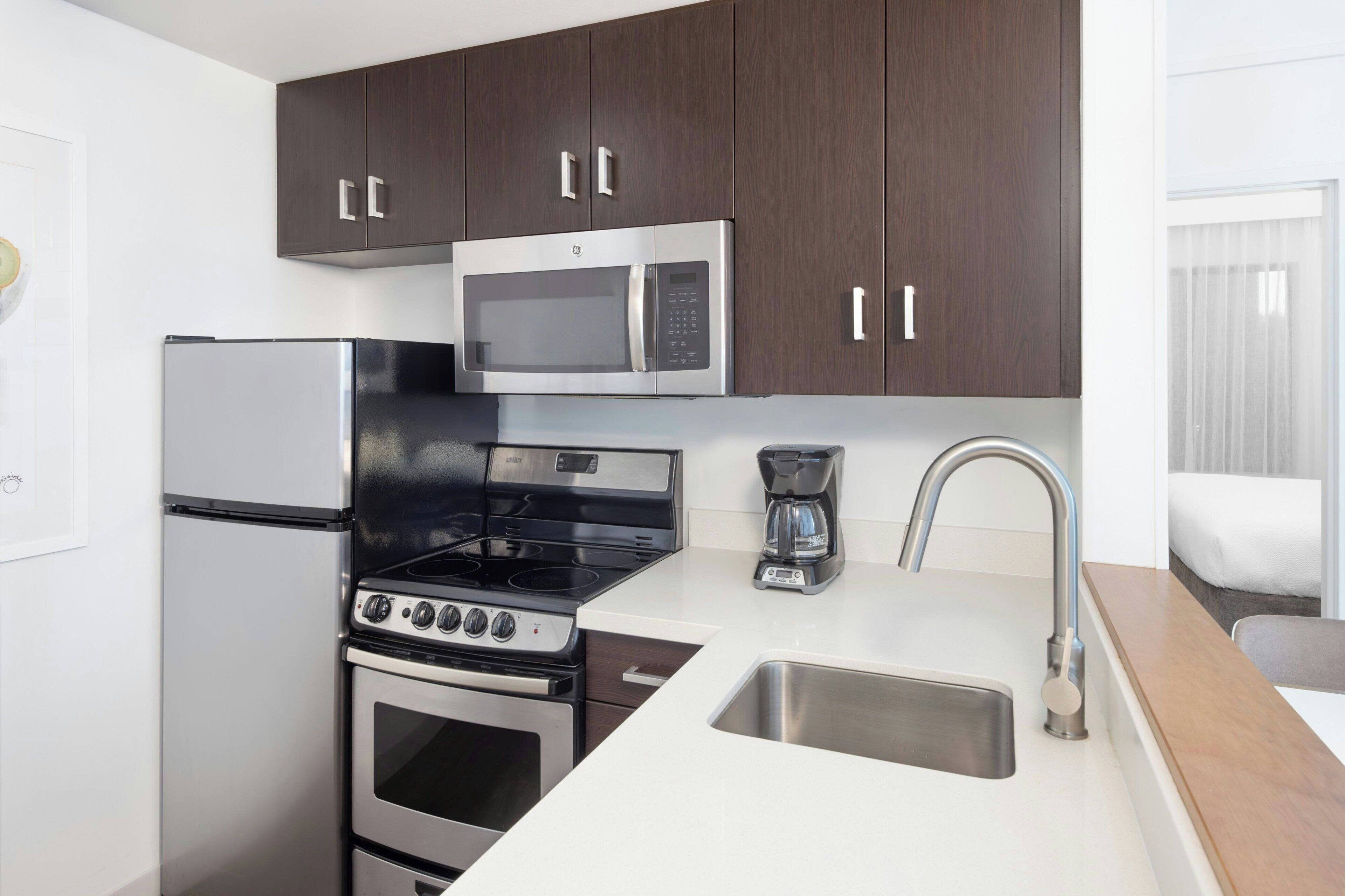 Una cocina compacta con armarios de madera oscura, un refrigerador de acero inoxidable, un microondas, un horno, una estufa, una cafetera y un lavamanos con un grifo moderno.