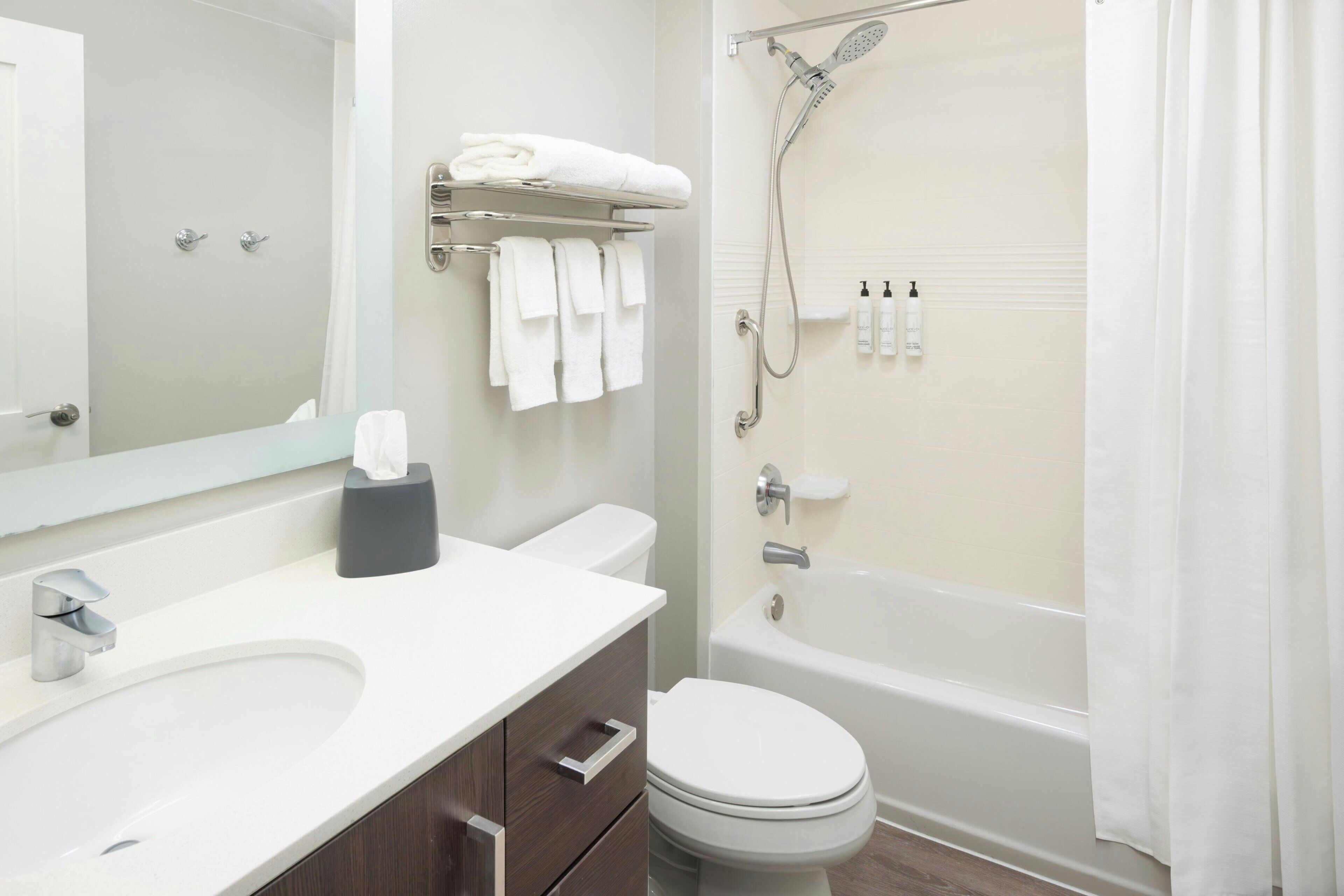 A modern bathroom with a white sink, vanity, and toilet. There is a shower with a handheld showerhead and a glass door. Towel racks and toiletries are visible.