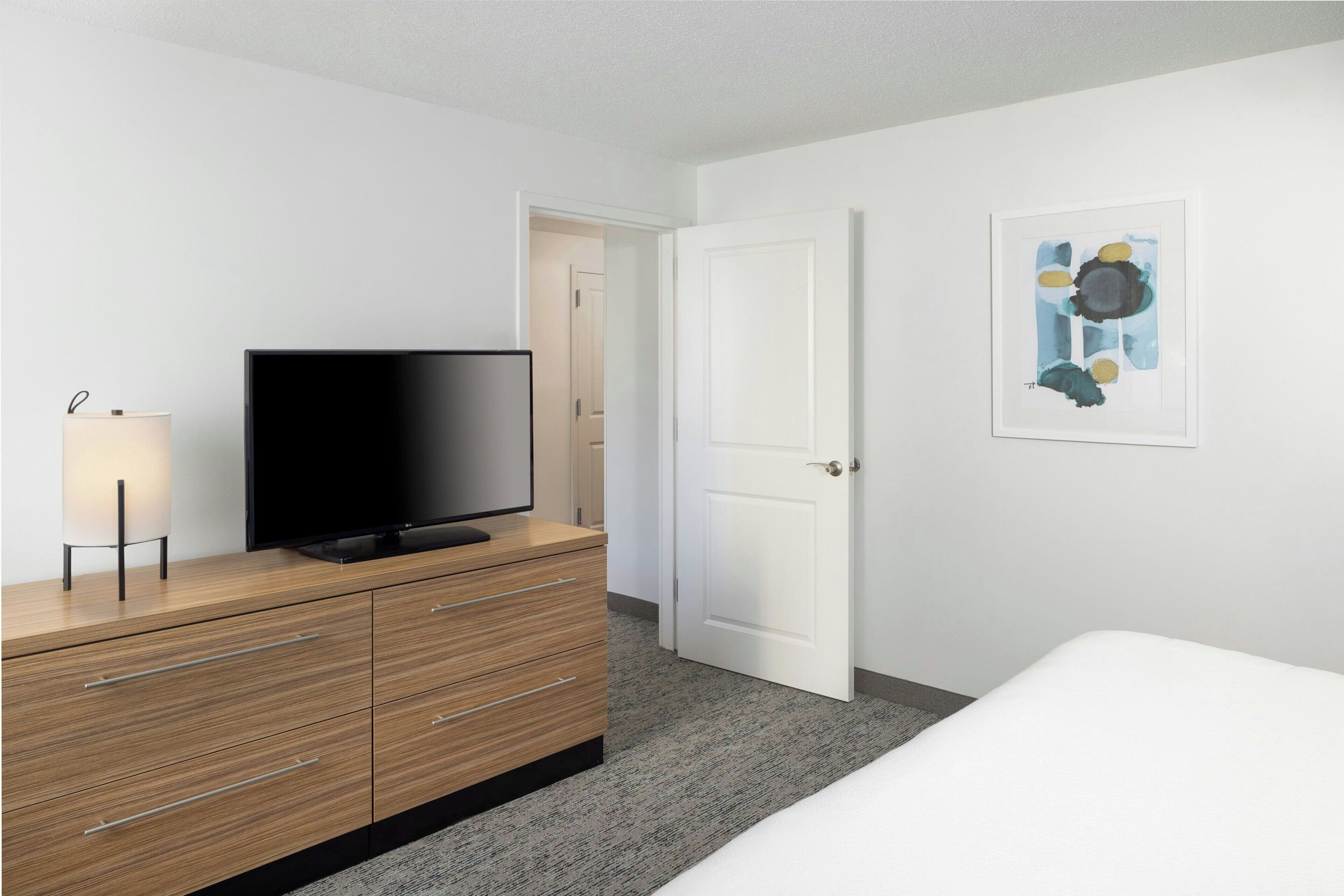 Habitación de hotel con una televisión de pantalla plana sobre un tocador de madera, una lámpara de noche, una puerta y un cuadro abstracto enmarcado en la pared.