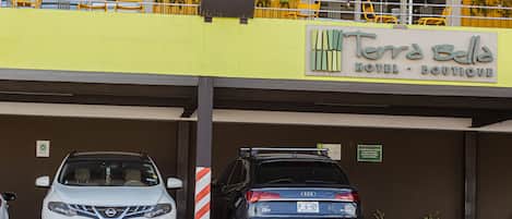 A parking area with two cars, a yellow building with a sign reading "Terra Bella Hotel - Boutique," and a balcony with yellow chairs.