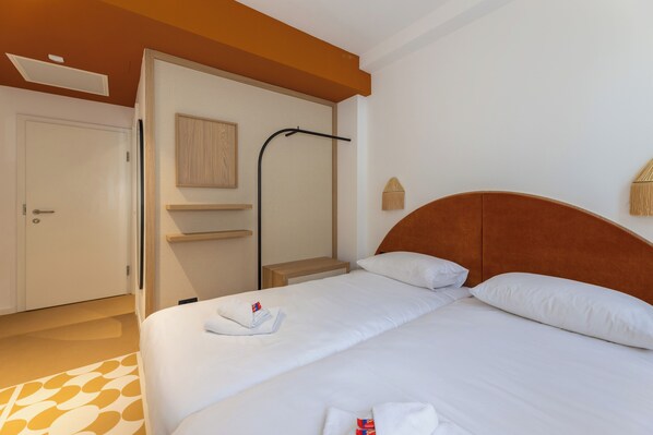 A hotel room with a bed, a door, a towel, a nightstand, and a wall-mounted light fixture.