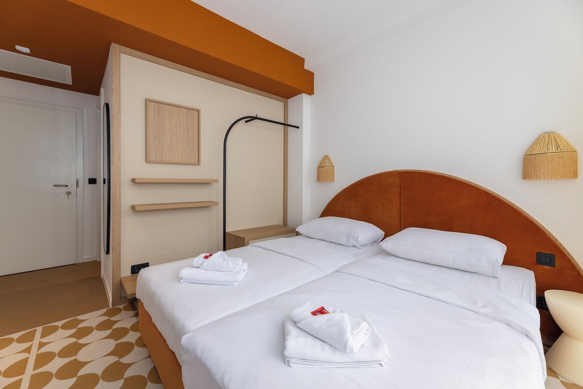 A hotel room with two beds, a wooden headboard, a white door, a towel rack, and a wall-mounted light fixture.