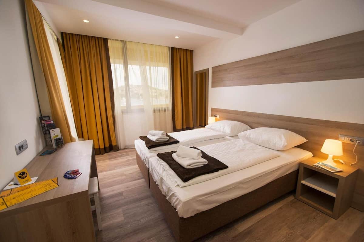 A hotel room with two beds, a desk with a chair, a nightstand with a lamp, and a window with curtains.