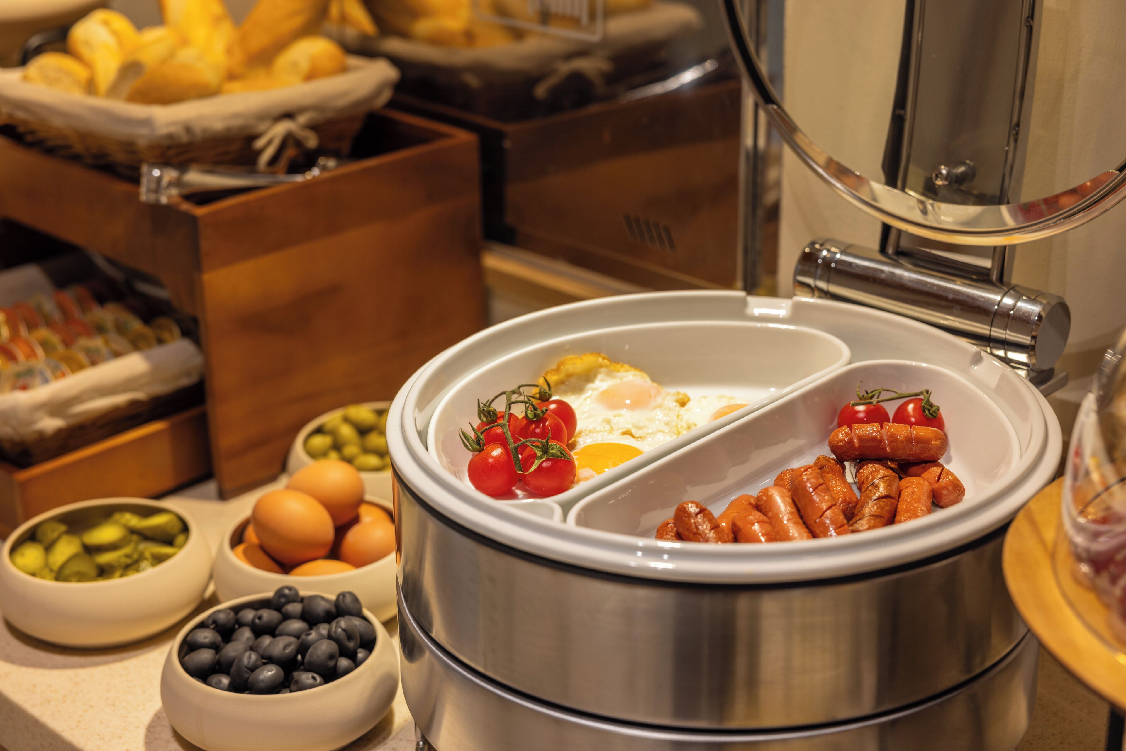 Buffet com diversos alimentos, incluindo ovos, linguiça, tomates e azeitonas.