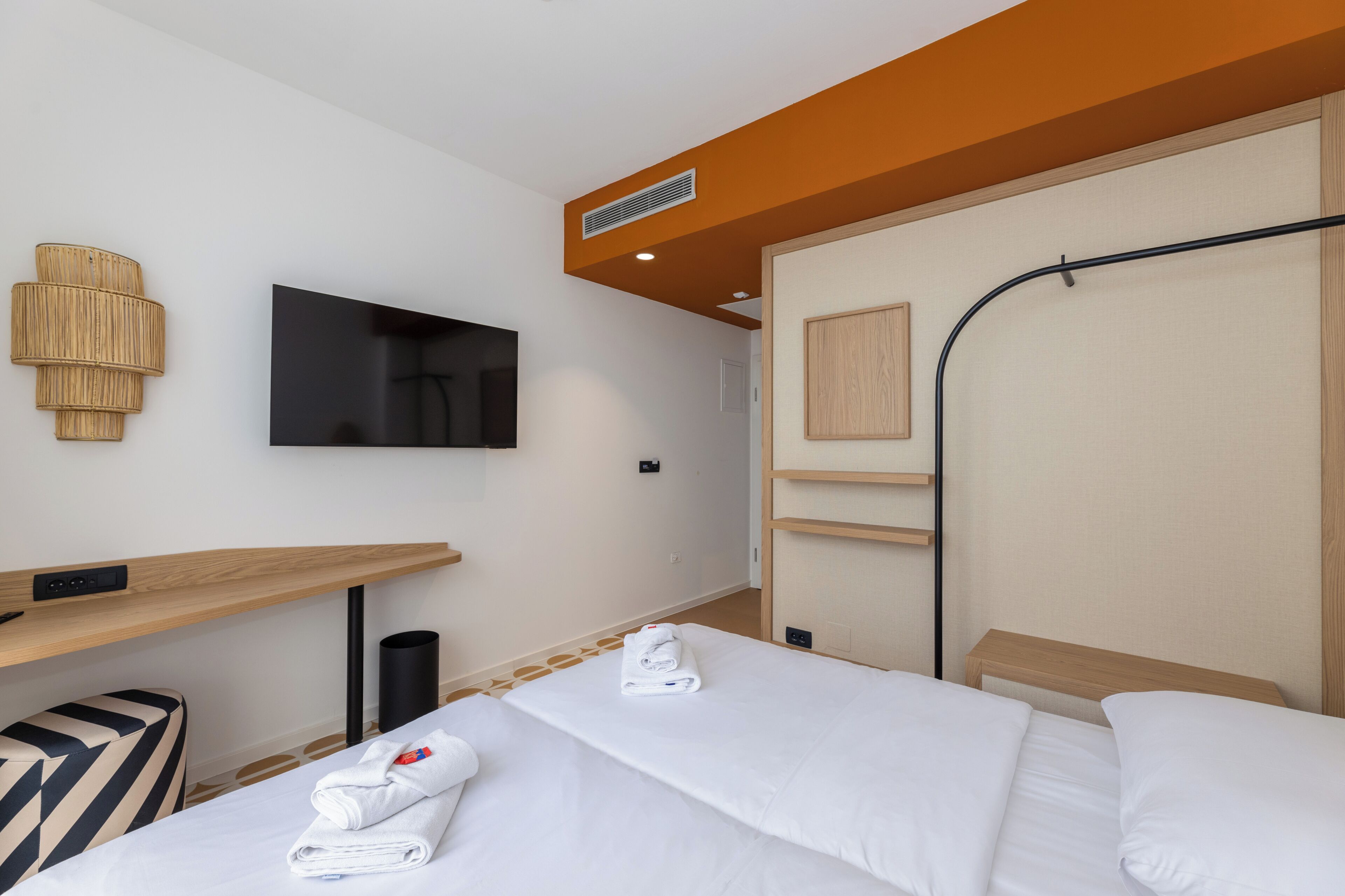 A hotel room with a bed, a TV, a desk, and a wall-mounted lamp.